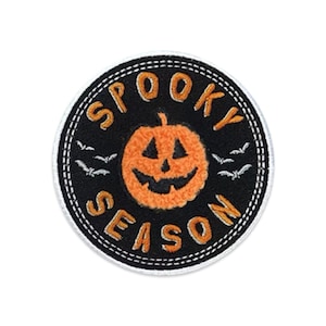 May include: A circular embroidered patch with a black background and a white border. The center features an orange jack-o'-lantern with a smiling face, surrounded by small white bats. The text SPOOKY SEASON is stitched in orange letters.