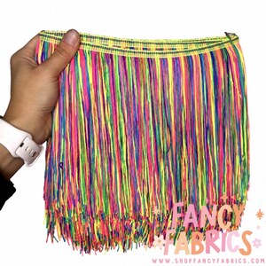 Puede incluir: Un fleco multicolor con hilos de colores neón amarillo, rosa, azul y verde que cuelgan de una cinta tejida. El adorno presenta una textura densa y superpuesta con una paleta de colores arcoíris. El texto FANCY FABRICS es visible.