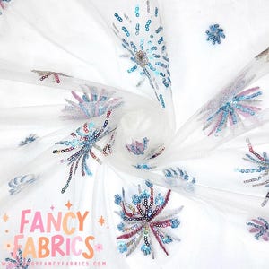 May include: White tulle fabric with a repeating pattern of sequined floral designs in shades of blue, pink, and silver. The fabric has a delicate, airy appearance. The text "FANCY FABRICS" is visible in the lower left corner.