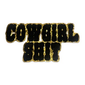 Puede incluir: Un parche bordado de dos líneas con el texto COWGIRL SHIT en letras negras y gruesas de estilo bloque. Cada letra está delineada con un borde grueso y brillante de color dorado que crea un efecto texturizado alrededor de la tela negra.