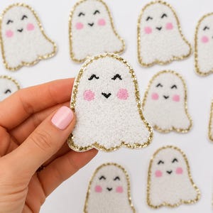 Puede incluir: Un parche con forma de fantasma blanco de superficie texturizada, con ojos y boca bordados en negro y mejillas rosadas. El parche está delineado con un borde de purpurina dorada, y varios parches de fantasmas idénticos se disponen al fondo.