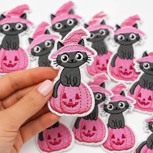May include: A collection of embroidered patches featuring a grey cat wearing a pink witch hat and sitting on a pink jack-o'-lantern pumpkin. The patches have a white border and are arranged in a group, with one patch held up in the foreground.