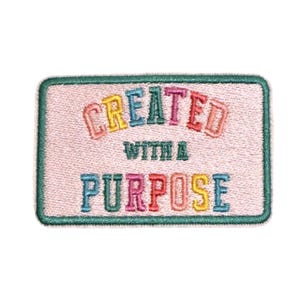 Pode incluir: Um emblema retangular bordado com fundo rosa claro e borda verde escura. O texto "CREATED WITH A PURPOSE" está costurado no centro com linha multicolorida, incluindo letras nas cores amarela, azul, verde, rosa e laranja.