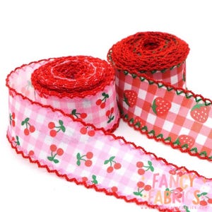May include: Two rolls of decorative ribbon with a gingham pattern. One ribbon features cherries, the other strawberries. Both ribbons have a red scalloped edge. The ribbons are white, pink, and red. The text "FANCY FABRICS" is visible.