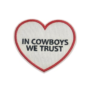 May include: A heart-shaped embroidered patch with a white fabric base and a red stitched border. The text IN COWBOYS WE TRUST is embroidered in black capital letters across the center of the heart, creating a clear and bold visual statement.