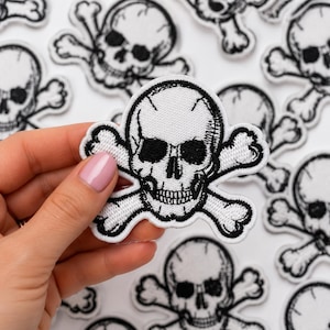 May include: A white embroidered patch featuring a black skull and crossbones design with detailed stitching. The patch has a clean white background and a thick black border, showcasing a classic pirate-inspired motif with high-contrast thread work.