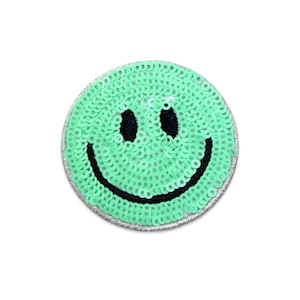 May include: A circular patch featuring a bright mint green sequin surface. The design includes two black embroidered oval eyes and a wide black embroidered curved mouth, creating a classic smiley face expression on a round fabric base.