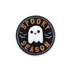 May include: A circular embroidered patch with a black background and a white border. The center features a white ghost with a smiling face, surrounded by small white bats. The orange text SPOOKY SEASON is arched around the ghost.