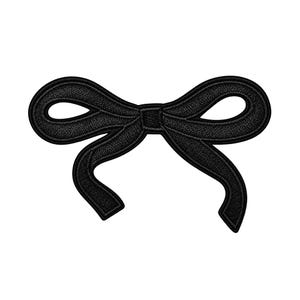May include: A black embroidered patch in the shape of a bow. The design features a central knot with two symmetrical loops and two trailing ribbon ends, all rendered with a dense, textured thread pattern against a solid black background.