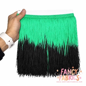 Puede incluir: Una tira de flecos con una sección superior de color verde vibrante que transiciona a una sección inferior de color negro sólido. El fleco está unido a una cinta de cabecera blanca. El texto FANCY FABRICS aparece en la esquina inferior.