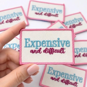 May include: A rectangular embroidered patch with a white background and a pink stitched border. The text Expensive is embroidered in light blue, while the words and difficult are embroidered in a dark pink cursive script font.