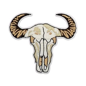 May include: An embroidered patch featuring a bull skull with large, curved horns. The skull is rendered in shades of cream, beige, and white, while the horns are detailed with dark brown and tan stitching, all outlined in a white border.