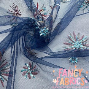 May include: Navy blue tulle fabric with embroidered floral designs in shades of teal, pink, and burgundy. The fabric has a delicate, sheer appearance, with the text "FANCY FABRICS" in the lower right corner.