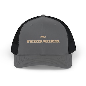 May include: A charcoal gray trucker hat with a black mesh back and a solid front panel. The front features a small tan graphic of a fish above the tan text "WHISKER WARRIOR" in a bold, blocky font. The hat has a curved brim and a button on top.