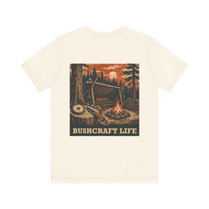 Könnte beinhalten: Ein cremefarbenes T-Shirt mit einer rechteckigen Grafik auf der Rückseite. Die Illustration zeigt ein Waldlager mit einer Schutzhütte, einem Lagerfeuer, einem Kompass und einem Messer auf einem Baumstumpf. Der Text BUSHCRAFT LIFE steht darunter.