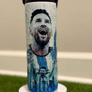 May include: A cylindrical white tumbler featuring a graphic print of a soccer player with a beard, shown in multiple poses wearing a blue and white striped jersey. The design includes blue paint splatter accents and a black lid with a flip-top spout.