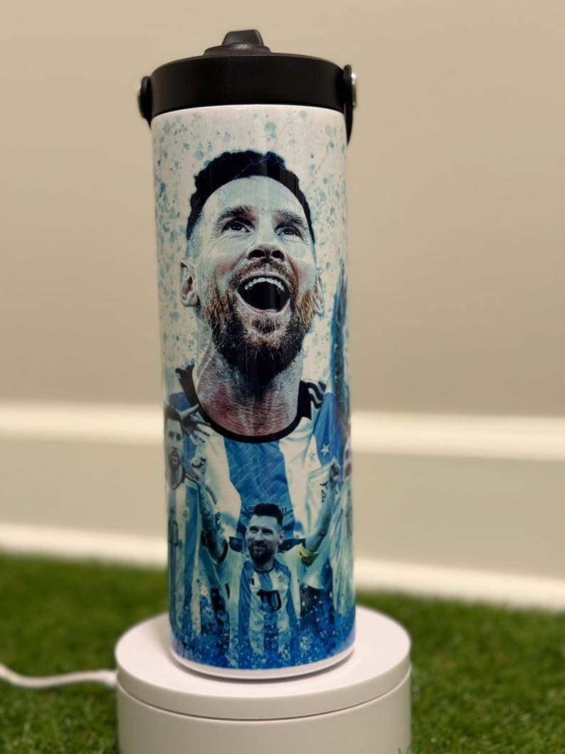 May include: A cylindrical white tumbler featuring a graphic print of a soccer player with a beard, shown in multiple poses wearing a blue and white striped jersey. The design includes blue paint splatter accents and a black lid with a flip-top spout.