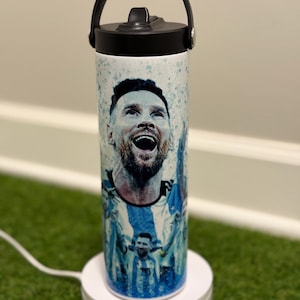 May include: A white cylindrical water bottle featuring a graphic print of a soccer player in a blue and white jersey. The bottle has a black lid with a carry handle and a straw spout. The design includes blue paint splatter accents.