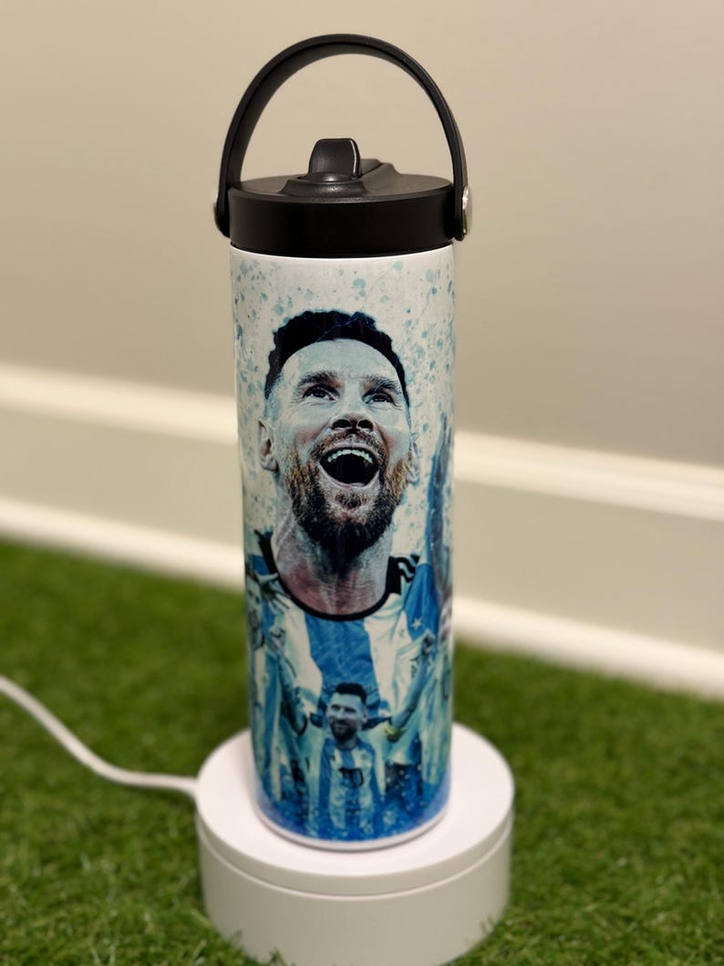 May include: A white cylindrical water bottle featuring a graphic print of a soccer player in a blue and white jersey. The bottle has a black lid with a carry handle and a straw spout. The design includes blue paint splatter accents.