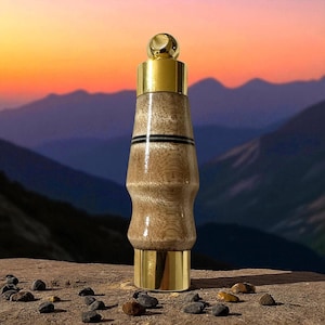 May include: A decorative bottle stopper with a gold-colored top and base. The body is made of light brown wood with a natural grain pattern and two black stripes. The stopper is standing on a stone surface.