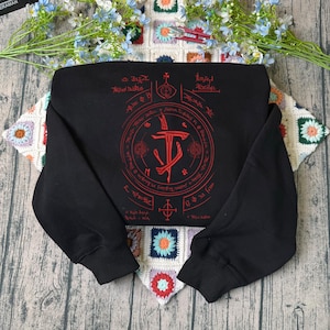 May include: Black sweatshirt with a red graphic design. The design features a central emblem with a sword and a circular pattern with text. The sweatshirt has long sleeves and a crew neck.