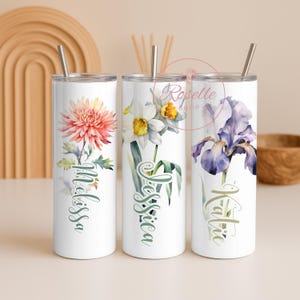 Può includere: Tre bicchieri bianchi in acciaio inossidabile con disegni floreali e nomi. Uno presenta una dalia rosa e arancione con il nome "Melissa". Un altro ha un narciso bianco con "Jessica". Il terzo ha un iris viola con "Katia". Ognuno ha una cannuccia d'argento.