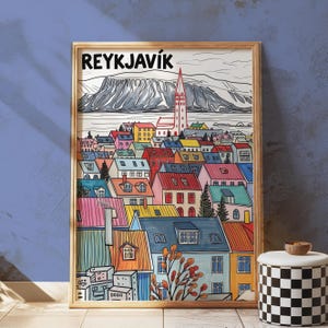 May include: A framed art print of Reykjavik, Iceland, featuring colorful rooftops and a tall church steeple. The artwork has a mountain range in the background and the word "REYKJAVIK" at the top. The print is in a wooden frame.