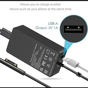 May include: A black rectangular AC power adapter with a proprietary connector cable and a built-in USB-A port. Text on the adapter includes 'Replacement AC Adapter' and 'USB-A Output: 5V 1A'. The device allows charging another device simultaneously.