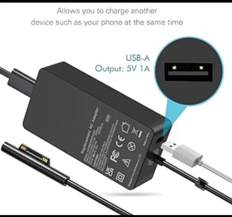May include: A black rectangular AC power adapter with a proprietary connector cable and a built-in USB-A port. Text on the adapter includes 'Replacement AC Adapter' and 'USB-A Output: 5V 1A'. The device allows charging another device simultaneously.