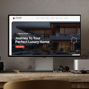 Op de afbeelding: Een computermonitor toont de website van Pillar Real Estate Solutions. De website toont een modern luxe huis met de tekst "Journey To Your Perfect Luxury Home". De monitor staat op een houten bureau met een toetsenbord, muis en dennenappels.