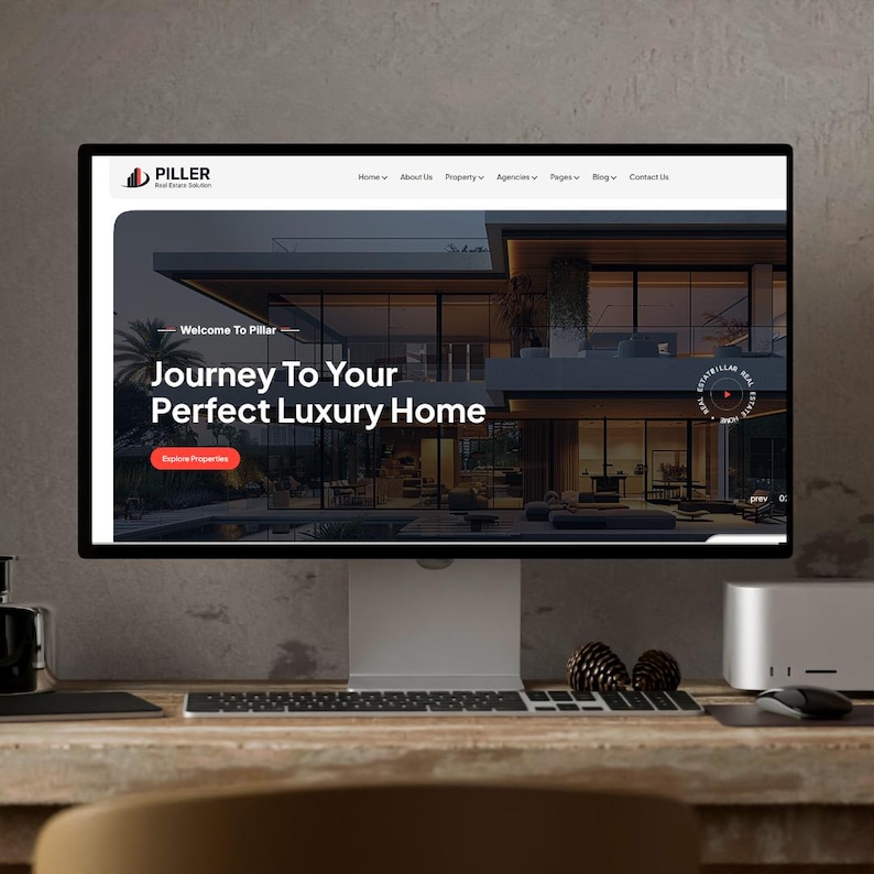 Op de afbeelding: Een computermonitor toont de website van Pillar Real Estate Solutions. De website toont een modern luxe huis met de tekst "Journey To Your Perfect Luxury Home". De monitor staat op een houten bureau met een toetsenbord, muis en dennenappels.