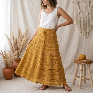 Puede incluir: Una falda larga de ganchillo color amarillo mostaza con cintura de cordón y patrones calados intrincados en toda la tela. La falda tiene un dobladillo festoneado y se combina con una sencilla camiseta blanca sin mangas y sandalias marrones.