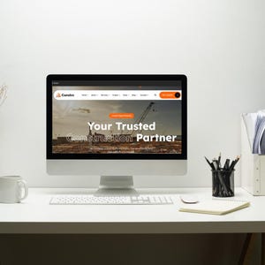 May include: A desktop computer displaying a website with the text "Your Trusted Construction Partner." The computer sits on a white desk with a keyboard, mouse, notepad, pencils, and a coffee mug. The website features a construction site image.