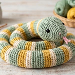 Könnte beinhalten: Eine geringelte Häkelschlange mit horizontalen Streifen in Salbeigrün, Senfgelb und Creme. Das Stofftier hat zwei schwarze Sicherheitsaugen aus Kunststoff und eine kleine, rosafarbene, gespaltene Zunge, die aus dem Mund herausragt.