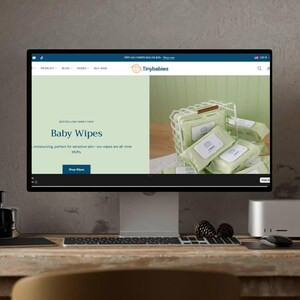 May include: A computer monitor displays a website for "Tinybabies" selling baby wipes. The screen shows packages of baby wipes in a wire basket. The website text includes "Baby Wipes" and "Shop Wipes". The monitor sits on a wooden desk.