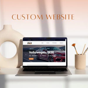 May include: A laptop displaying a website with the text "CUSTOM WEBSITE." The website features a red Volkswagen, 1938 model. A beige vase and a small container with paintbrushes are on the table.