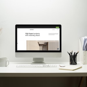 May include: A desktop computer displays a website with the text "High-Impact projects with visionary result." The computer sits on a white desk with a keyboard, mouse, mug, notepad, and pencil holder. The background is a plain white wall.