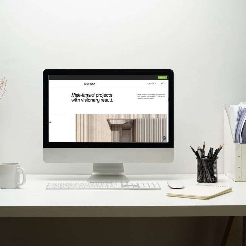 May include: A desktop computer displays a website with the text "High-Impact projects with visionary result." The computer sits on a white desk with a keyboard, mouse, mug, notepad, and pencil holder. The background is a plain white wall.