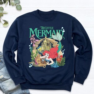 Może przedstawiać: Granatowa bluza z nadrukiem Arielki z Małej Syrenki. Projekt zawiera Ariel, Florka, Sebastiana i napis "The Little Mermaid" w kolorze zielonym. Bluza ma okrągły dekolt i długie rękawy.
