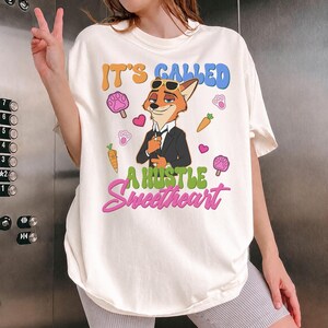 May include: Cream-colored t-shirt featuring a cartoon fox in a suit with sunglasses. The shirt has the text "IT'S CALLED A HUSTLE Sweetheart" in colorful bubble letters, along with paw prints, hearts, and carrots.