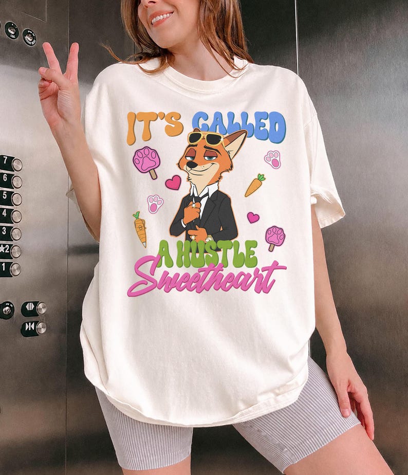 May include: Cream-colored t-shirt featuring a cartoon fox in a suit with sunglasses. The shirt has the text "IT'S CALLED A HUSTLE Sweetheart" in colorful bubble letters, along with paw prints, hearts, and carrots.