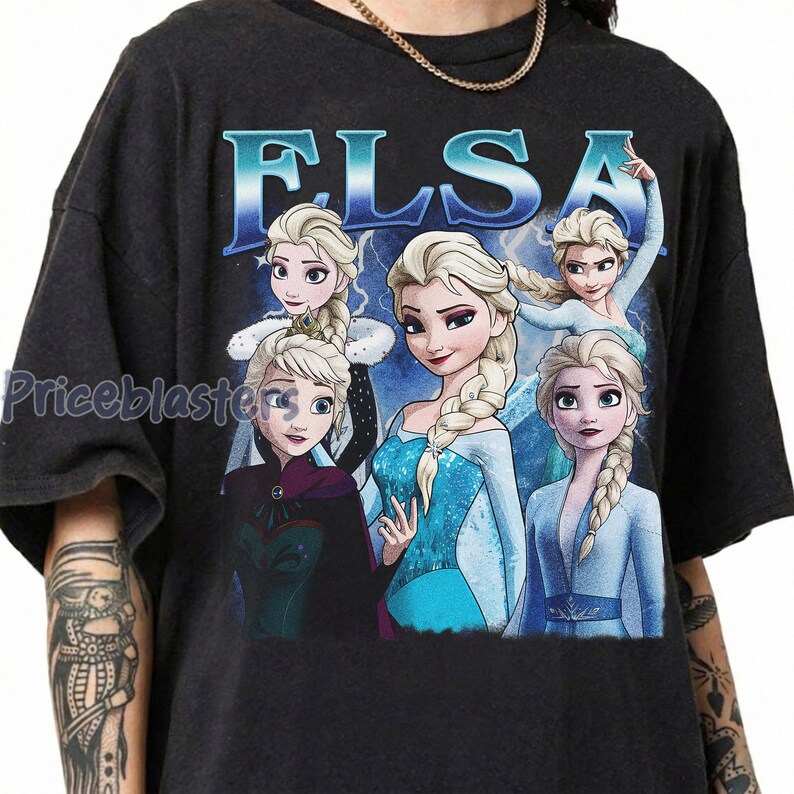 May include: Dark gray t-shirt featuring the name "ELSA" in blue letters above a graphic of the character Elsa from the movie Frozen. The graphic shows Elsa in various poses with a blue and white background.