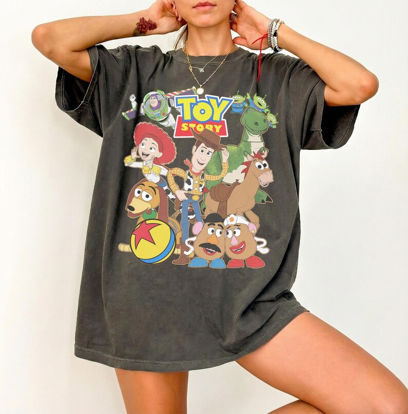 May include: Dark gray t-shirt featuring a colorful graphic of characters from the animated film "Toy Story." The design includes Woody, Buzz Lightyear, Jessie, and other characters. The shirt has a vintage, worn look.