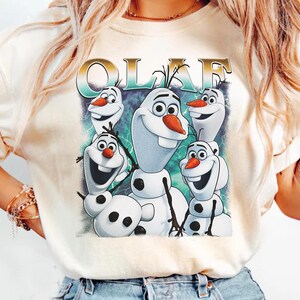 Puede incluir: Camiseta color crema con un gr&aacute;fico de Olaf de Frozen. El dise&ntilde;o incluye m&uacute;ltiples personajes de Olaf con caras sonrientes, narices de zanahoria y brazos de palo. La palabra "OLAF" est&aacute; impresa encima de los personajes en oro y verde azulado.