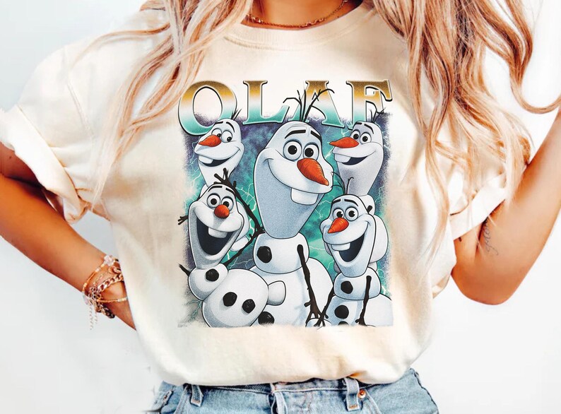 Puede incluir: Camiseta color crema con un gr&aacute;fico de Olaf de Frozen. El dise&ntilde;o incluye m&uacute;ltiples personajes de Olaf con caras sonrientes, narices de zanahoria y brazos de palo. La palabra "OLAF" est&aacute; impresa encima de los personajes en oro y verde azulado.