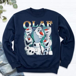 Puede incluir: Sudadera azul marino con un dise&ntilde;o de Olaf de Frozen. El dise&ntilde;o incluye m&uacute;ltiples personajes de Olaf con narices de zanahoria y la palabra "OLAF" en una fuente degradada. La sudadera est&aacute; hecha de un material suave.