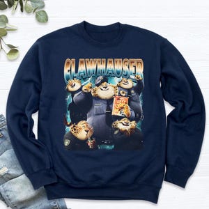 Puede incluir: Sudadera azul marino con un dise&ntilde;o de Clawhauser de Zootopia. El dise&ntilde;o muestra a Clawhauser con uniforme de polic&iacute;a, sosteniendo una bolsa de Lucky Chomps. La palabra "CLAWHAUSER" est&aacute; impresa encima de los personajes.