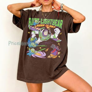 May include: Brown t-shirt featuring a graphic of Buzz Lightyear in a space scene with the text "Buzz Lightyear Lazer Blast". The design includes planets, stars, and other characters from the movie.