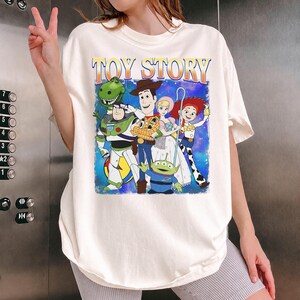Puede incluir: Camiseta color crema con un gr&aacute;fico de personajes de la pel&iacute;cula animada "Toy Story". El dise&ntilde;o incluye a Woody, Buzz Lightyear, Jessie y otros personajes. Las palabras "TOY STORY" est&aacute;n impresas en la parte superior en naranja y azul.