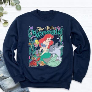 May include: Navy blue sweatshirt featuring a colorful graphic of Ariel from The Little Mermaid. The design includes Ariel, a dolphin, and other sea creatures, with the words "The Little Mermaid" in a teal font.